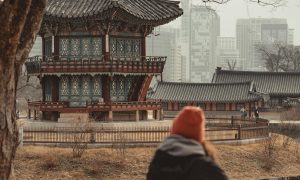 Back of a woman wearing woollen hat and jacket looking out to foreground of ornate building and backdrop of cityscape of skyrise buildings in South Korea.